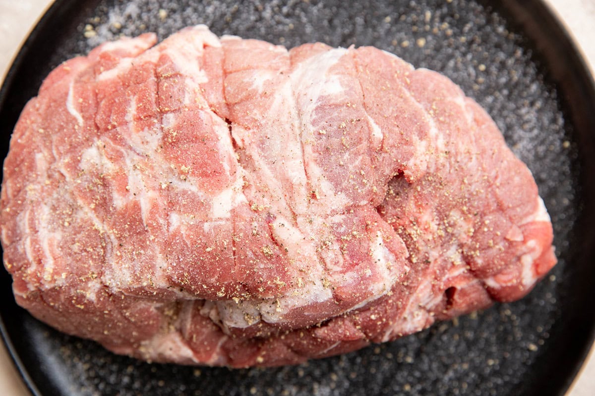 Pork butt roast seasoned with salt and pepper, sitting on a black plate.