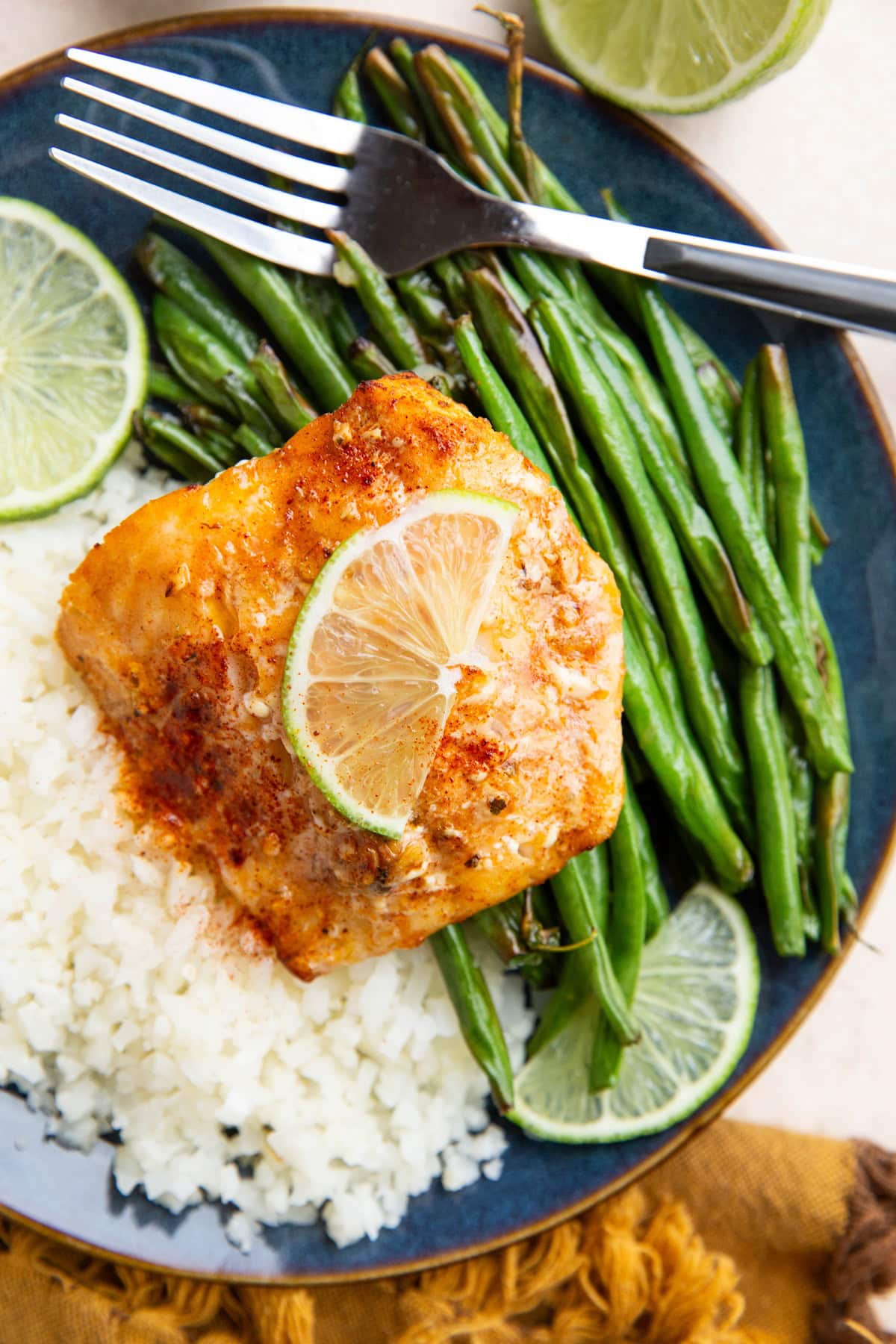 Cod on a blue plate with green beans and cauliflower rice and slices of limes.