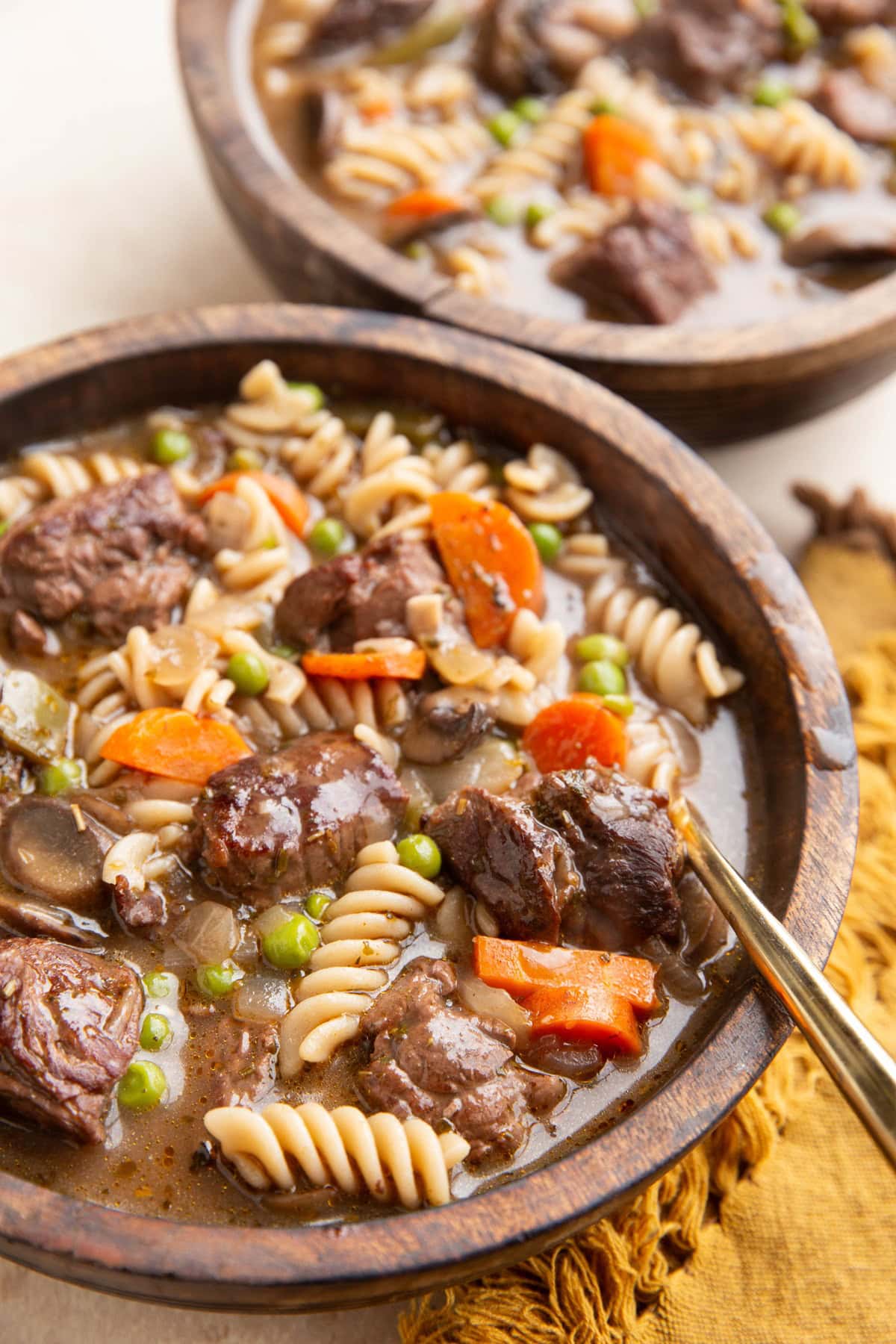 Two wooden bowls of beef noodle stew with a gold spoon and a gold napkin to the side.