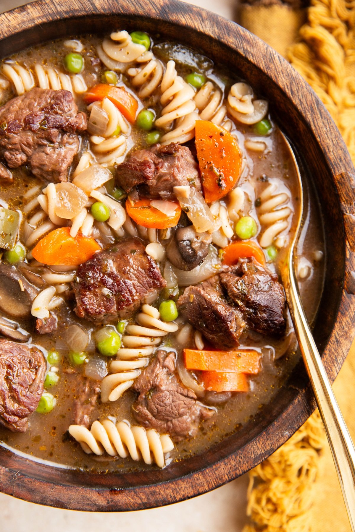 Wooden bowl of beef and noodle stew with a gold spoon and napkin to the side.