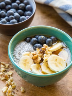 Teal bowl of chia pudding with sliced bananas, walnuts, and fresh berries on top. Chopped walnuts to the side, a bowl of fresh blueberries in the background, and a blue striped napkin.