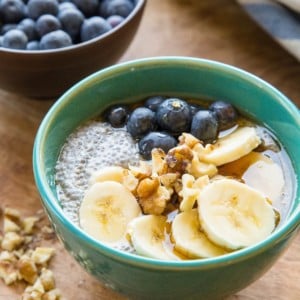 Teal bowl of chia pudding with sliced bananas, walnuts, and fresh berries on top. Chopped walnuts to the side, a bowl of fresh blueberries in the background, and a blue striped napkin.