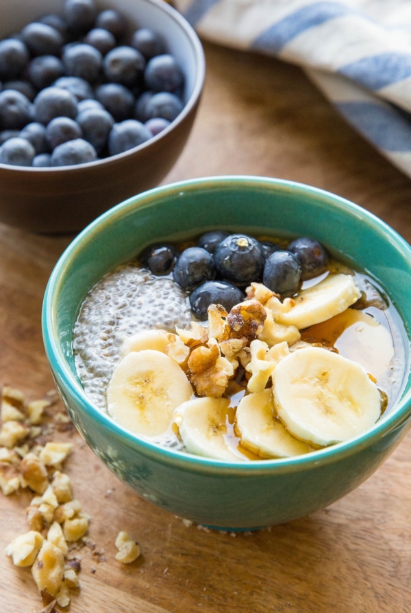 Teal bowl of chia pudding with sliced bananas, walnuts, and fresh berries on top. Chopped walnuts to the side, a bowl of fresh blueberries in the background, and a blue striped napkin.