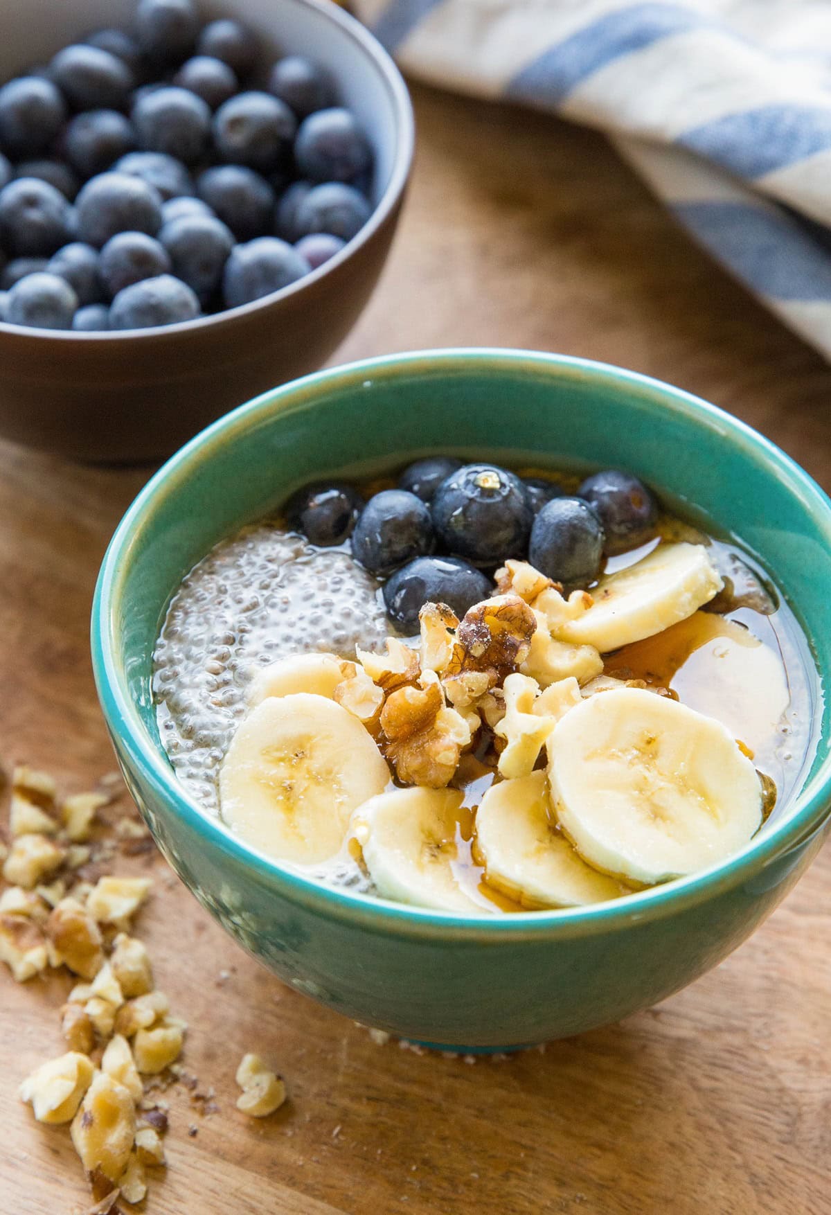 Teal bowl of chia pudding with sliced bananas, walnuts, and fresh berries on top. Chopped walnuts to the side, a bowl of fresh blueberries in the background, and a blue striped napkin.