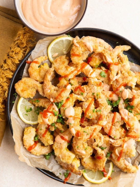 Black plate of pan-fried shrimp, drizzled in bang bang sauce with lemons to the side. A small bowl with more bang bang sauce and a napkin.