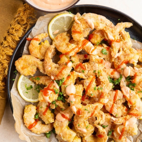 Black plate of pan-fried shrimp, drizzled in bang bang sauce with lemons to the side. A small bowl with more bang bang sauce and a napkin.