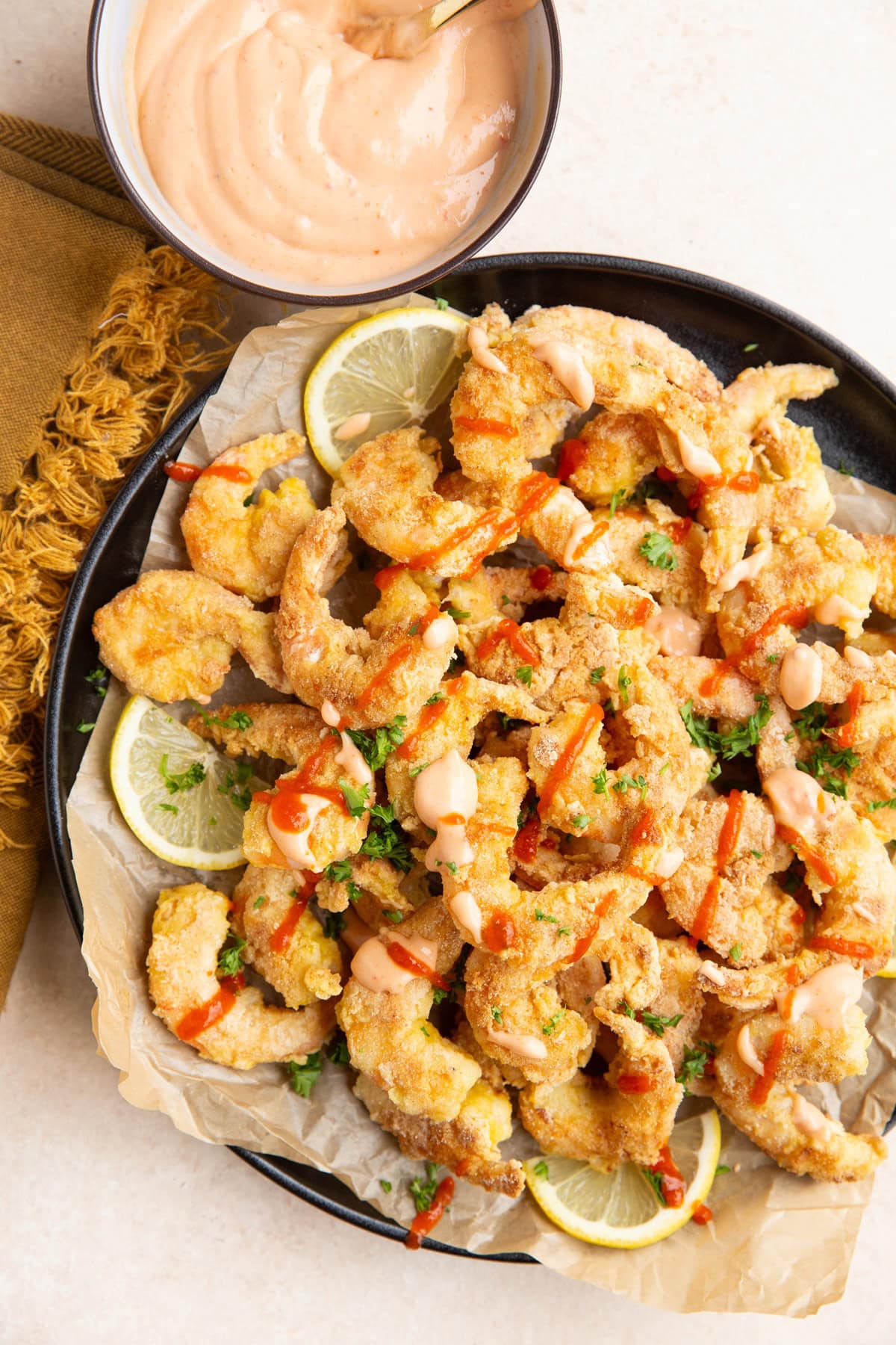 Black plate of pan-fried shrimp, drizzled in bang bang sauce with lemons to the side. A small bowl with more bang bang sauce and a napkin.