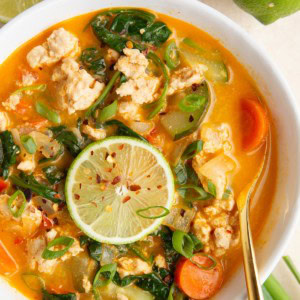 White bowl full of ground turkey curry soup with a bowl of red pepper flakes and fresh limes to the side.