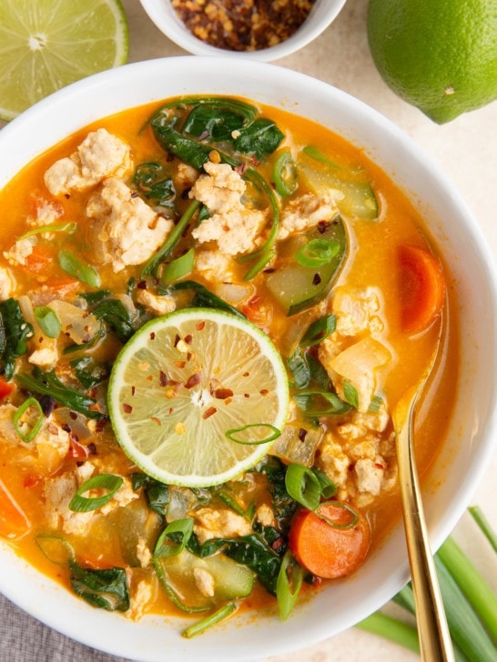 White bowl full of ground turkey curry soup with a bowl of red pepper flakes and fresh limes to the side.