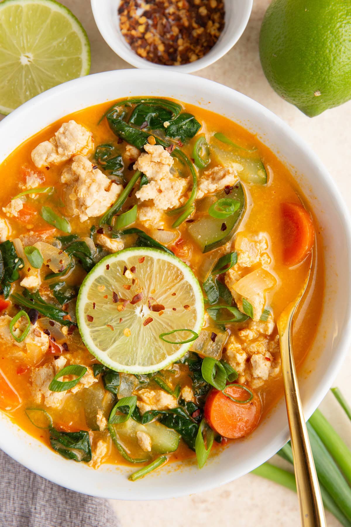 White bowl full of ground turkey curry soup with a bowl of red pepper flakes and fresh limes to the side.