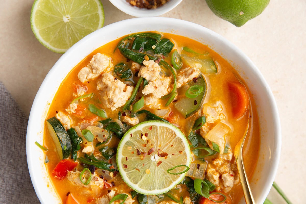 Bowl of Thai curry soup with a slice of lime and chopped green onions on top with a golden spoon and a grey napkin.