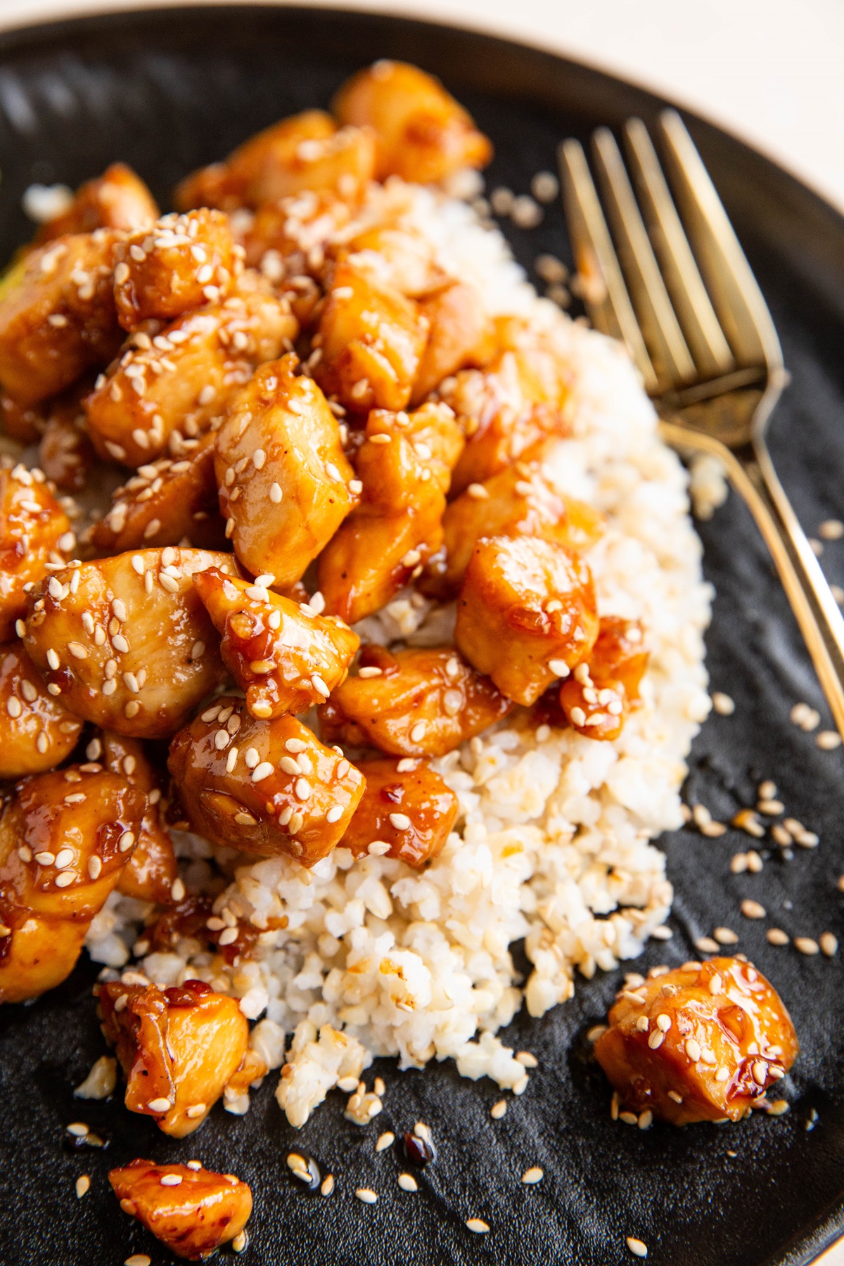 Black plate of brown rice with sesame chicken with a gold fork to the side.