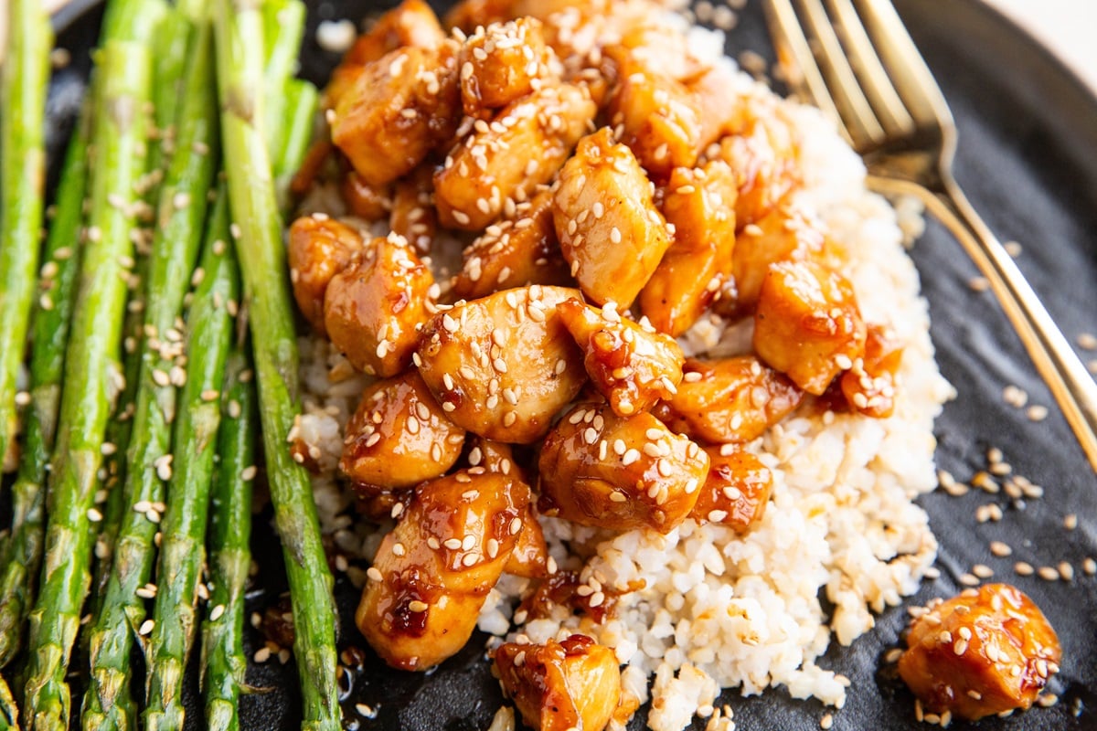 Black plate of brown rice, sesame chicken, and roasted asparagus.