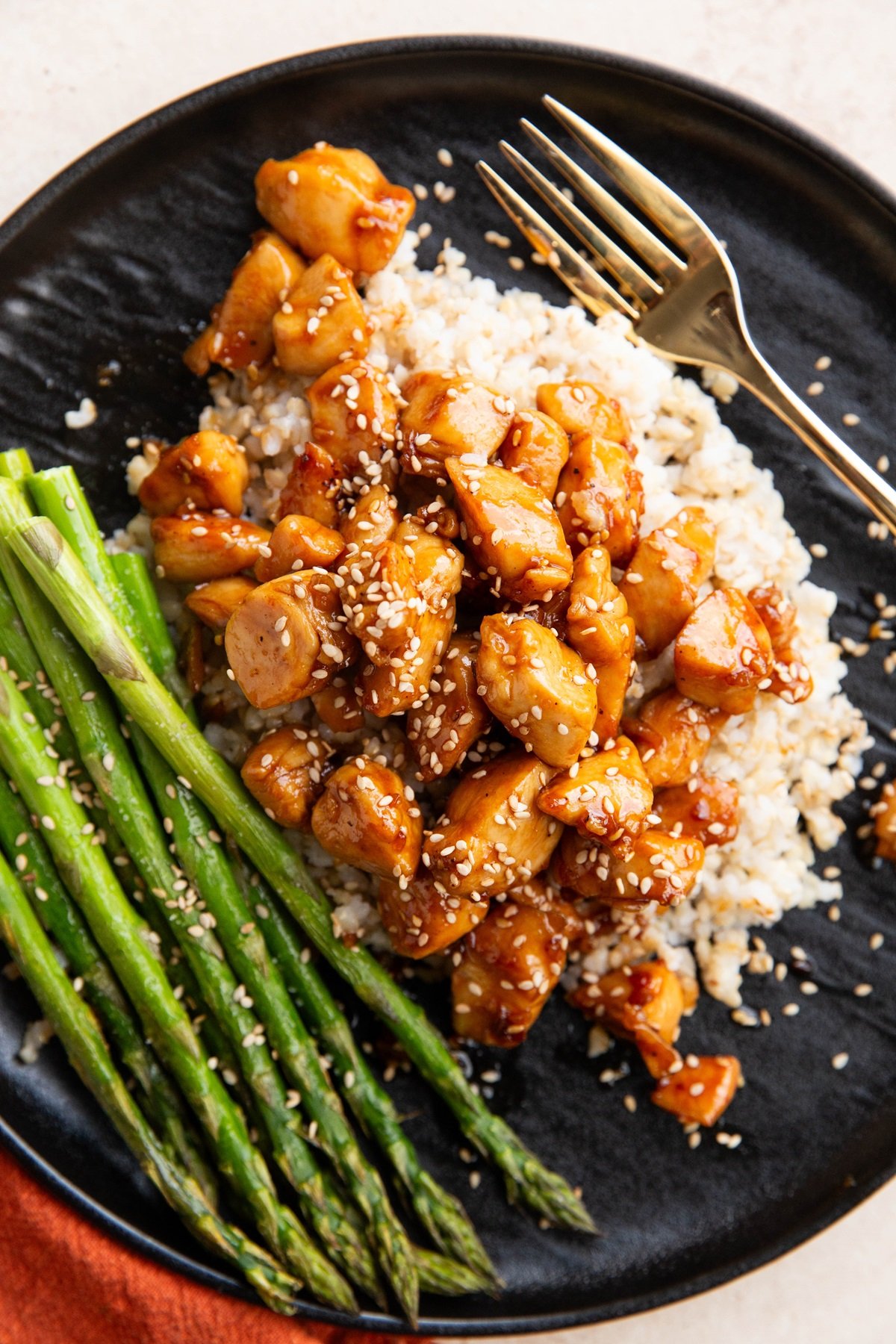 Black plate of sesame chicken with rice and asparagus. Red napkin and gold fork to the side.