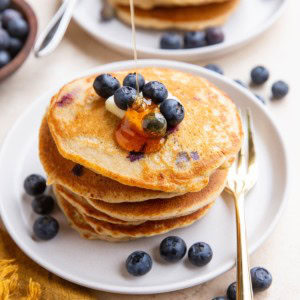 Honey being drizzled on a stack of blueberry oatmeal pancakes with blueberries and butter on top and a gold fork to the side.