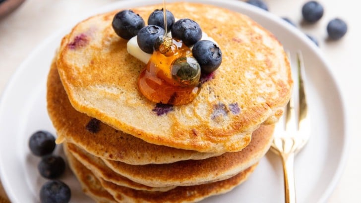 Honey being drizzled on a stack of blueberry oatmeal pancakes with blueberries and butter on top and a gold fork to the side.