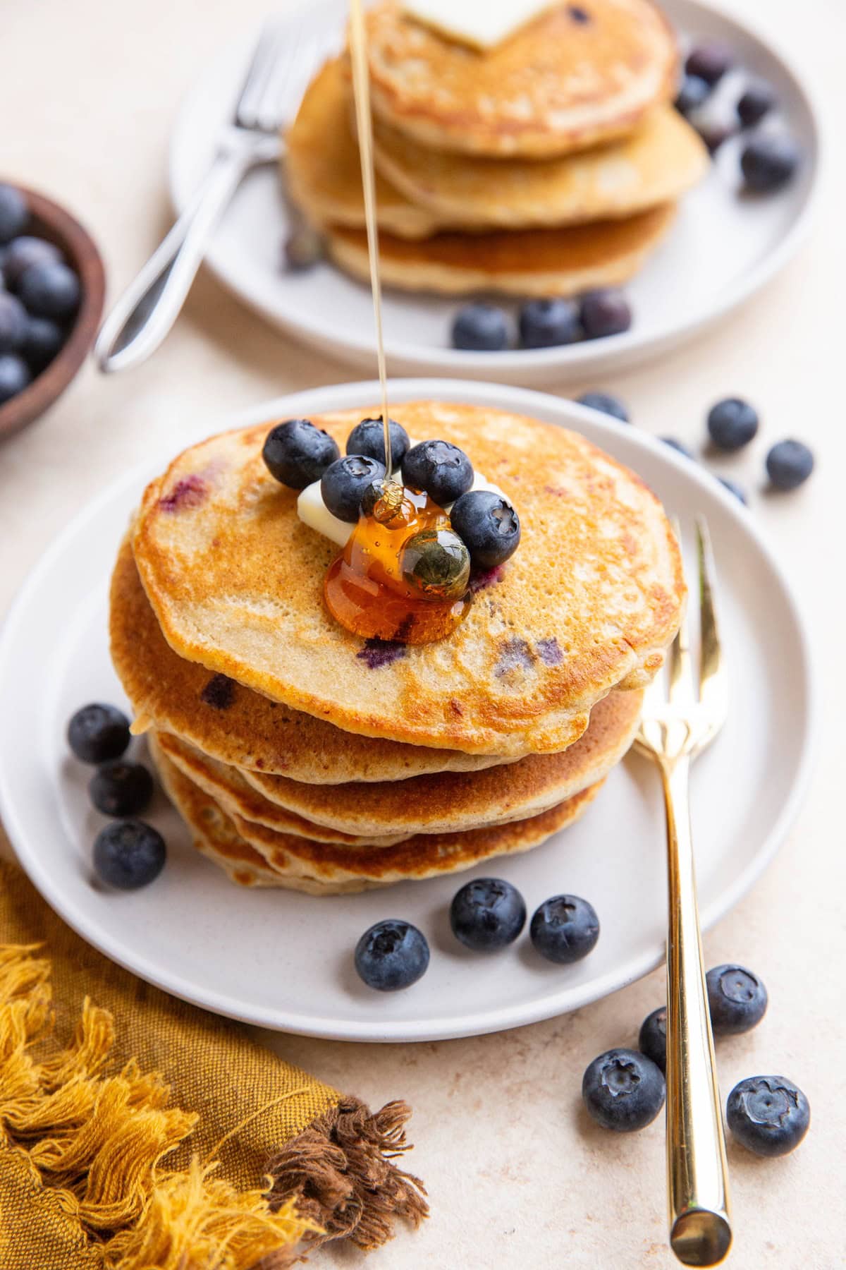 Honey being drizzled on a stack of blueberry oatmeal pancakes with blueberries and butter on top and a gold fork to the side.