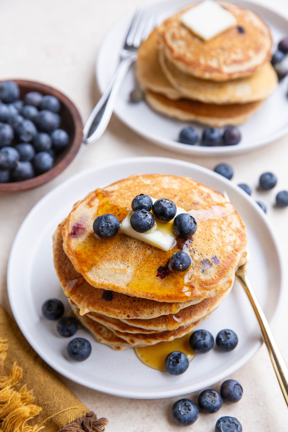 Two plates of blueberry pancakes with honey, butter, and fresh blueberries on top. A bowl of fresh blueberries to the side and a golden napkin.