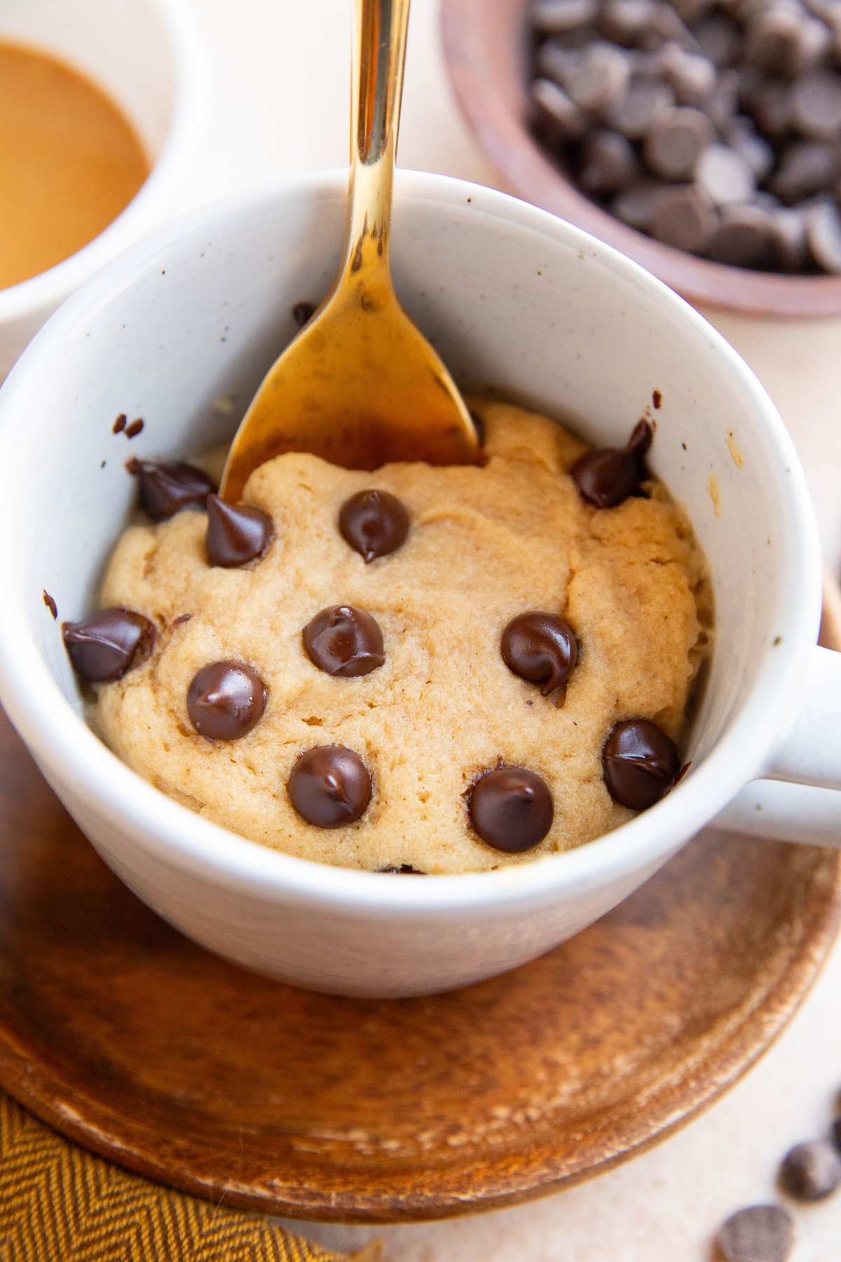 Peanut butter mug cake with chocolate chips on top and a gold spoon. Chocolate chips and peanut butter in the background.