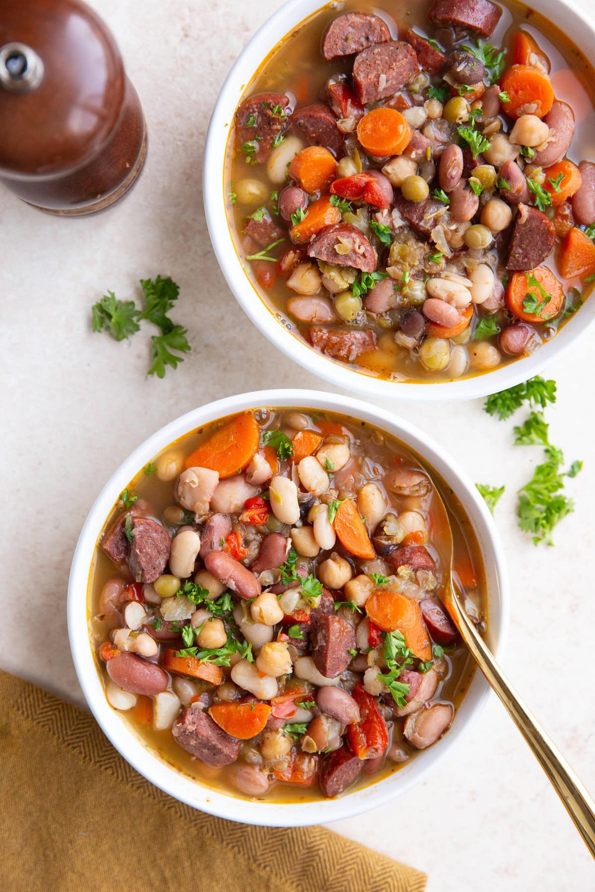 Two white bowls full of sausage and bean soup, ready to eat. pepper grinder, fresh parsley and napkin to the side.