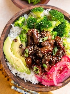 Wooden bowl of garlic steak with rice, pickled onions, broccoli and avocado. Chopsticks to the side and a golden napkin.