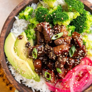 Wooden bowl of garlic steak with rice, pickled onions, broccoli and avocado. Chopsticks to the side and a golden napkin.