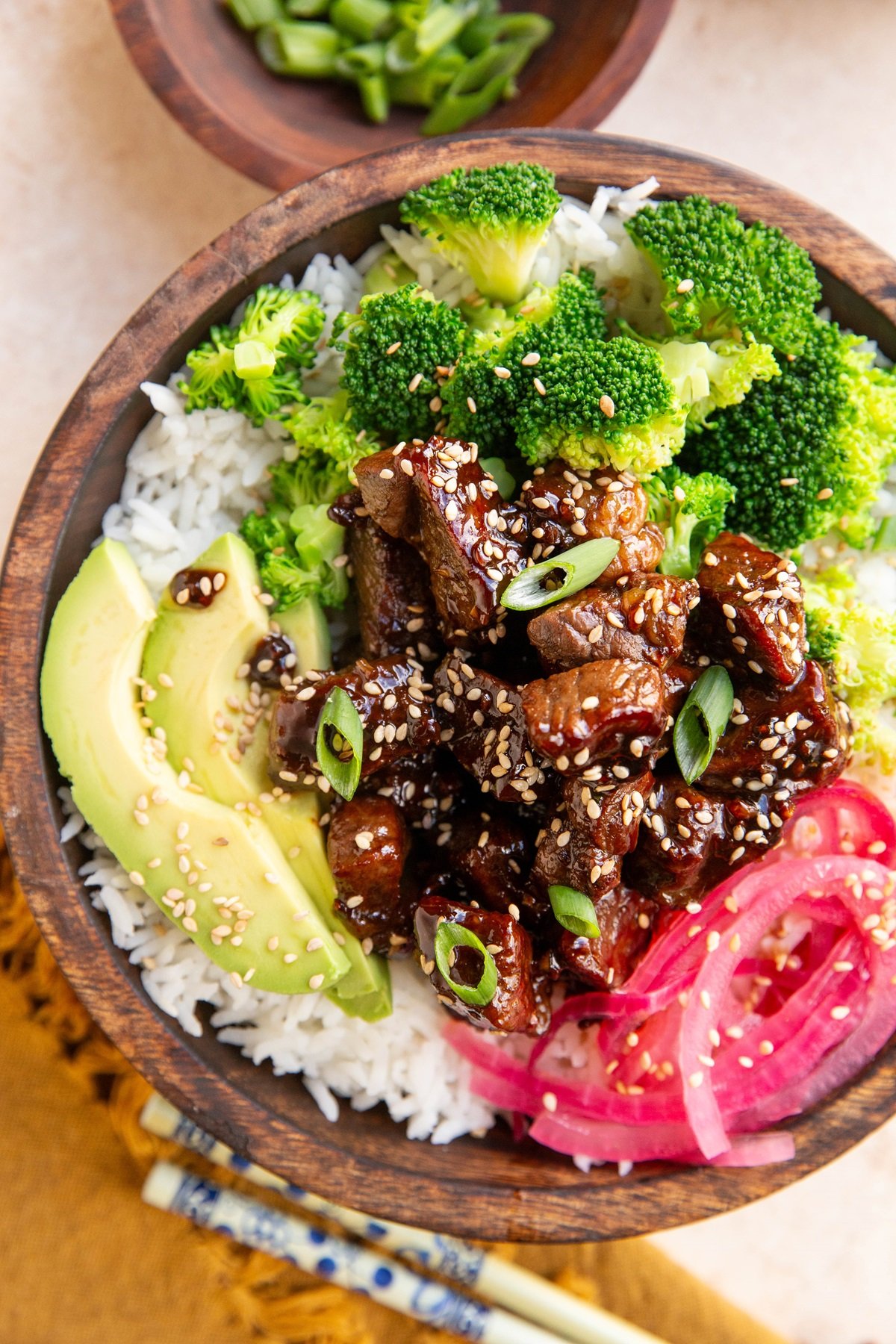 Wooden bowl of garlic steak with rice, pickled onions, broccoli and avocado. Chopsticks to the side and a golden napkin.