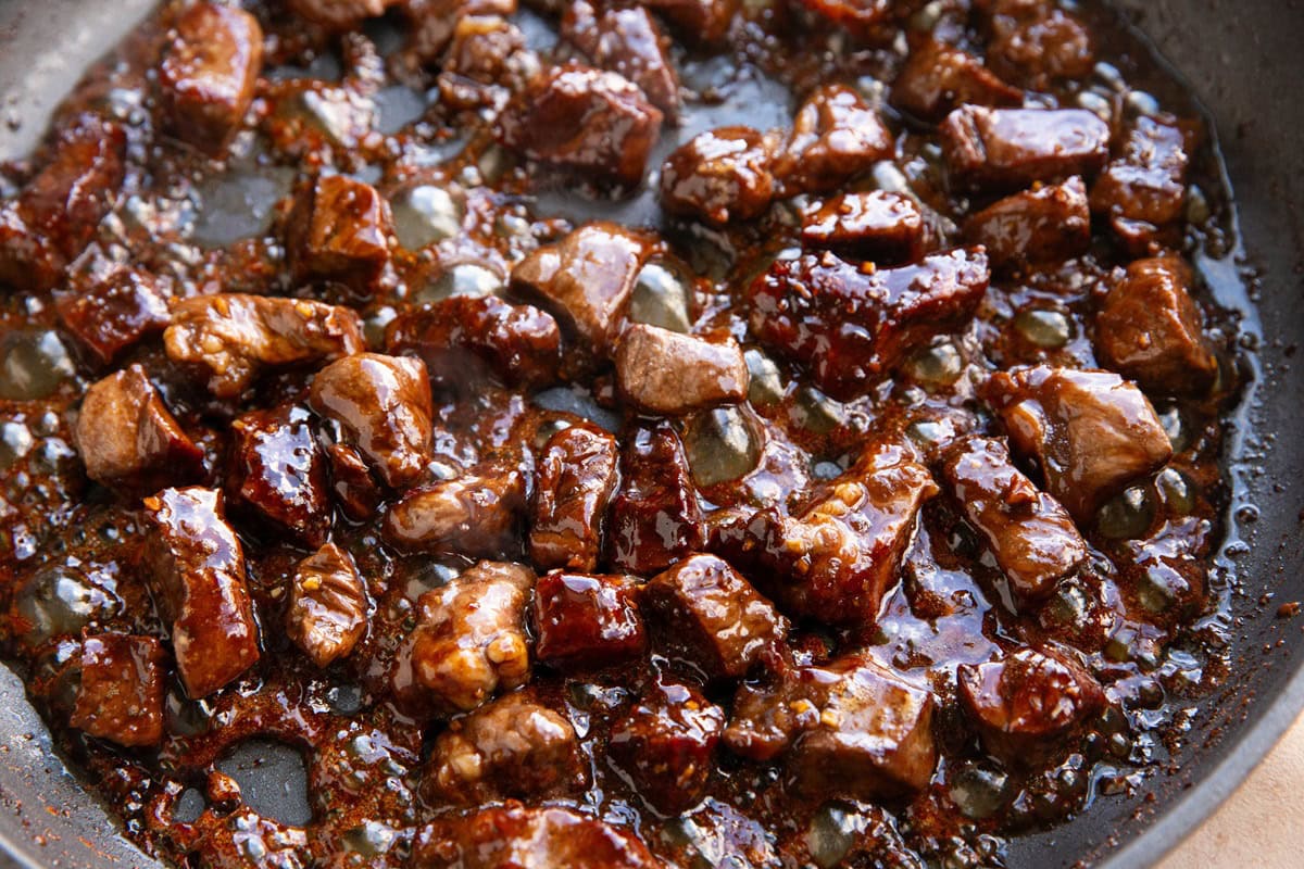 Steak bites cooking in a skillet in a thick and sticky sauce.