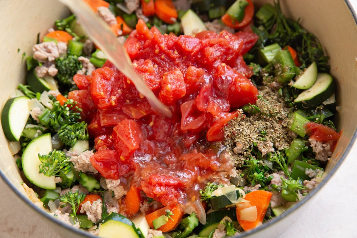 Diced tomatoes in a pot with veggies and ground turkey with chicken broth being poured in.