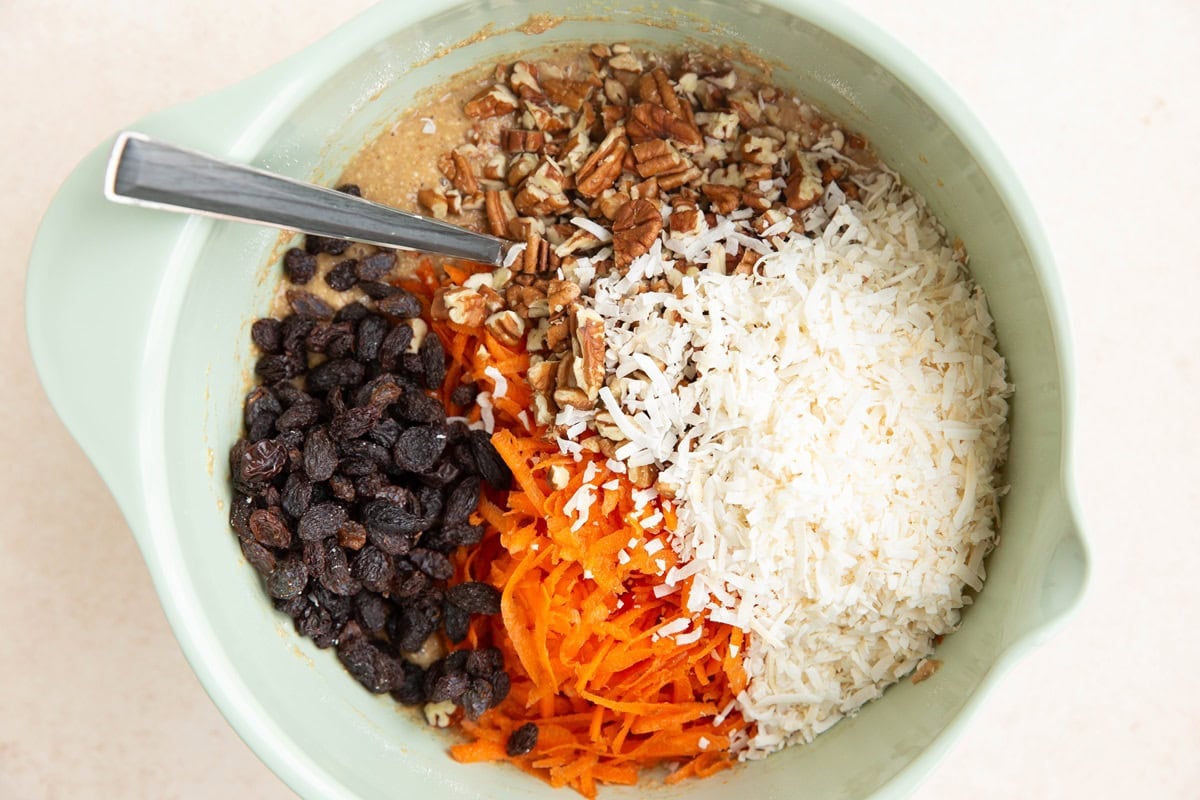 Mixing bowl of carrot cake batter with shredded carrot, shredded coconut, chopped pecans and raisins ready to be mixed in.