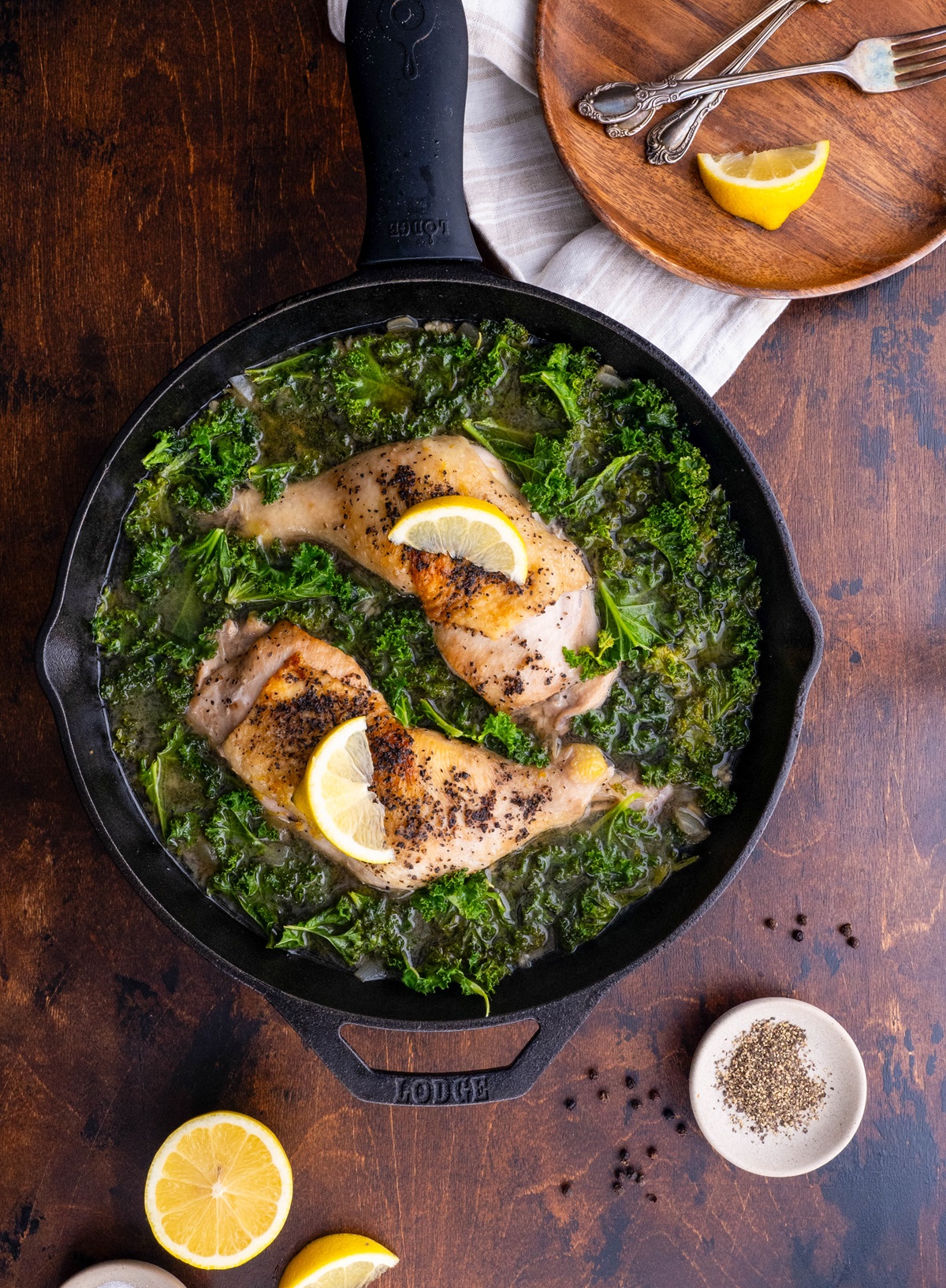 Large skillet with braised chicken and kale inside with a plate and lemons to the side, ready to serve.