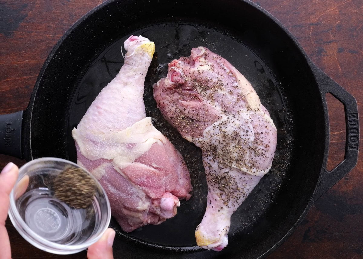 Cast iron skillet with chicken searing in it, being sprinkled with salt and pepper.