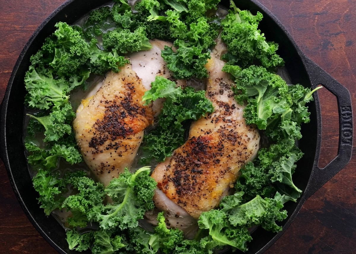 Skillet with chicken surrounded by raw kale leaves.