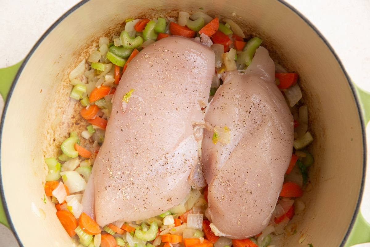 Two raw chicken breasts on top of sautéing vegetables in a Dutch oven.