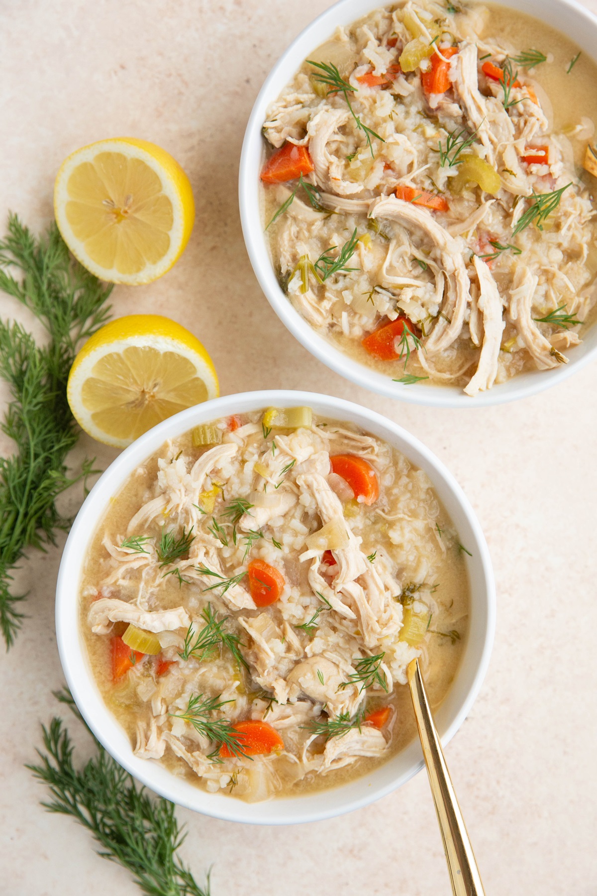 Two white bowls of Greek lemon chicken soup with fresh dill and cut lemons to the side.