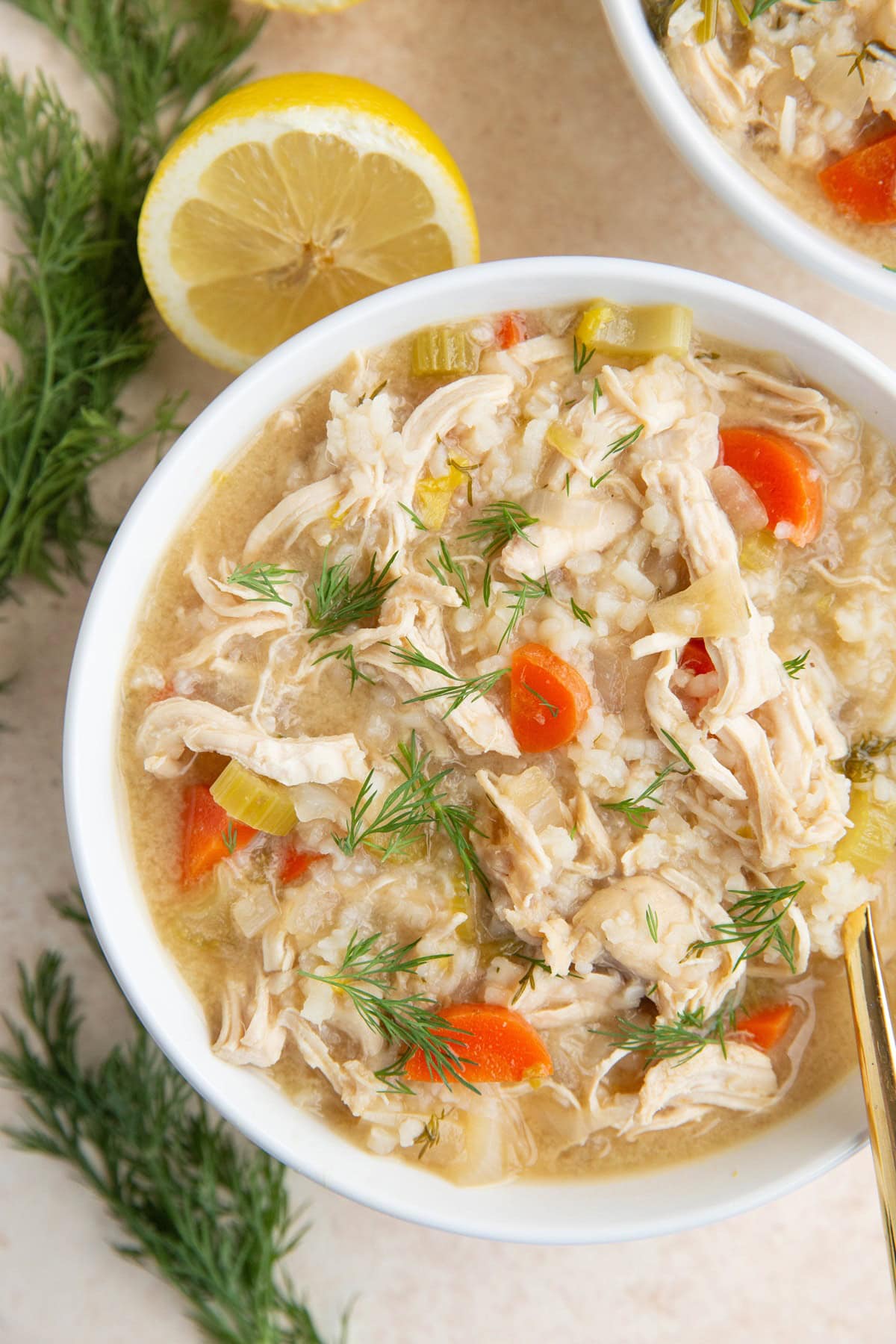 Big white bowl of lemon chicken soup with a gold spoon and fresh herbs around the bowl, ready to serve.