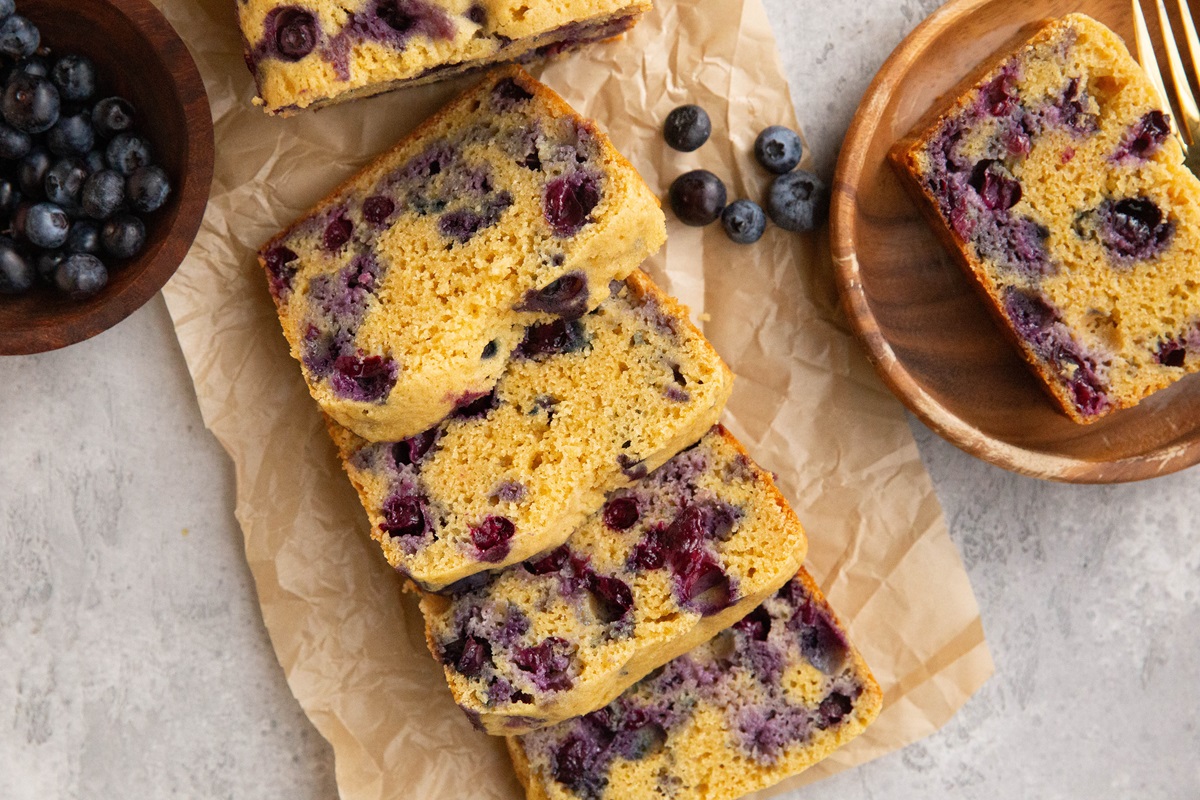 Almond flour blueberry bread sliced on a sheet of parchment paper with a slice of bread on a plate.
