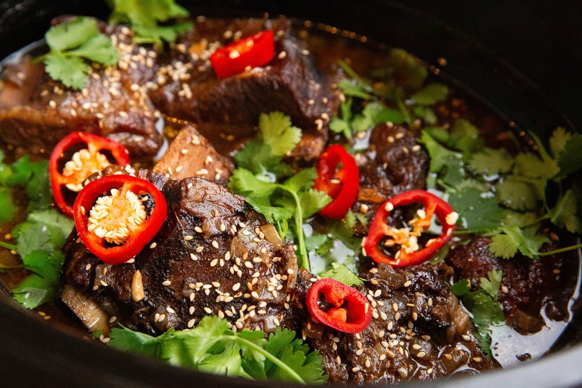 Asian short ribs recipe in a crock pot, sprinkled with sesame seeds, sliced red chilis, and fresh cilantro. Ready to serve.
