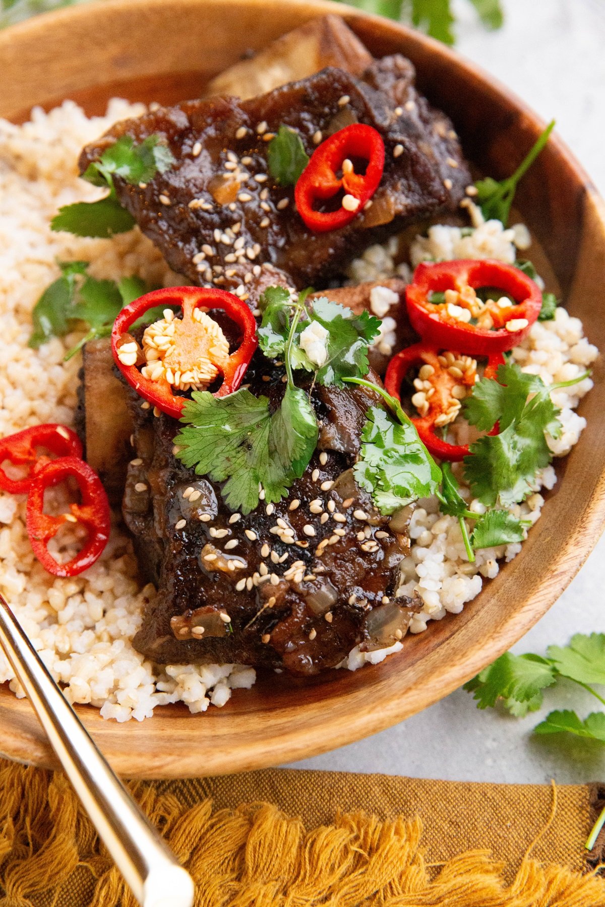 Wooden bowl with brown rice and Asian beef short ribs, sprinkled with sesame seeds, sliced red chilis, and fresh cilantro. with a golden fork and golden napkin to the side.