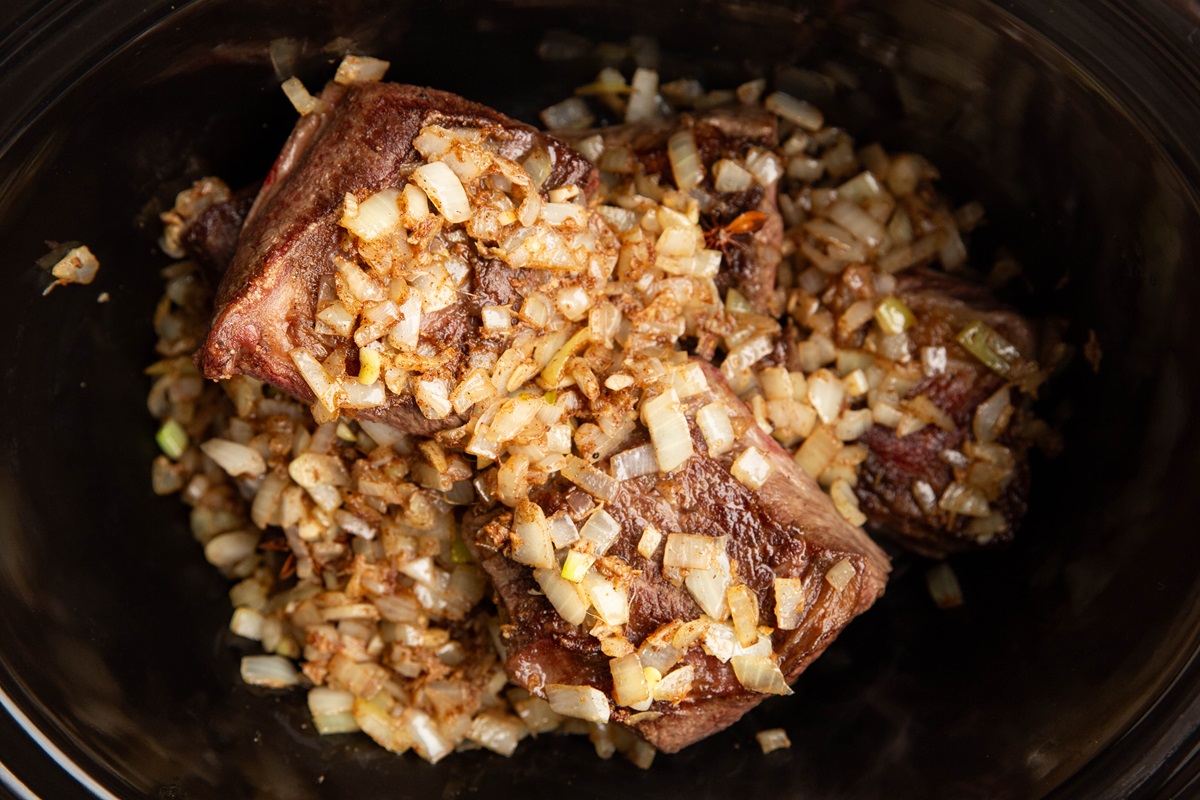Crock pot with seared short ribs and sauteed onions inside, ready to be made into Korean short ribs.