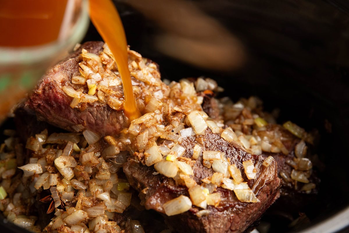 Pouring braising liquid into the slow cooker with the beef and onions.