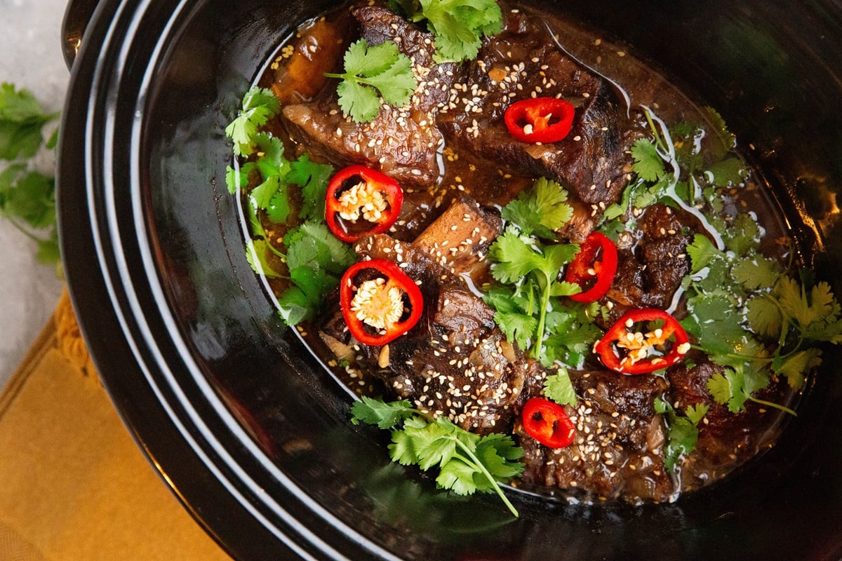 Crock pot with finished beef short ribs, ready to serve. Sprinkled with sesame seeds, sliced red chilis, and fresh cilantro.