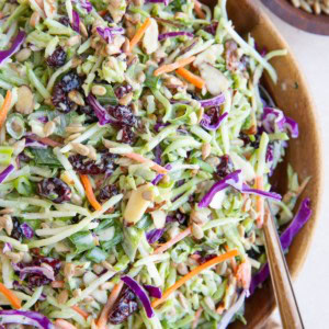 Wooden bowl full of broccoli slaw with a wood inlay spoon, ready to serve. A bowl of sunflower seeds to the side and a golden napkin.