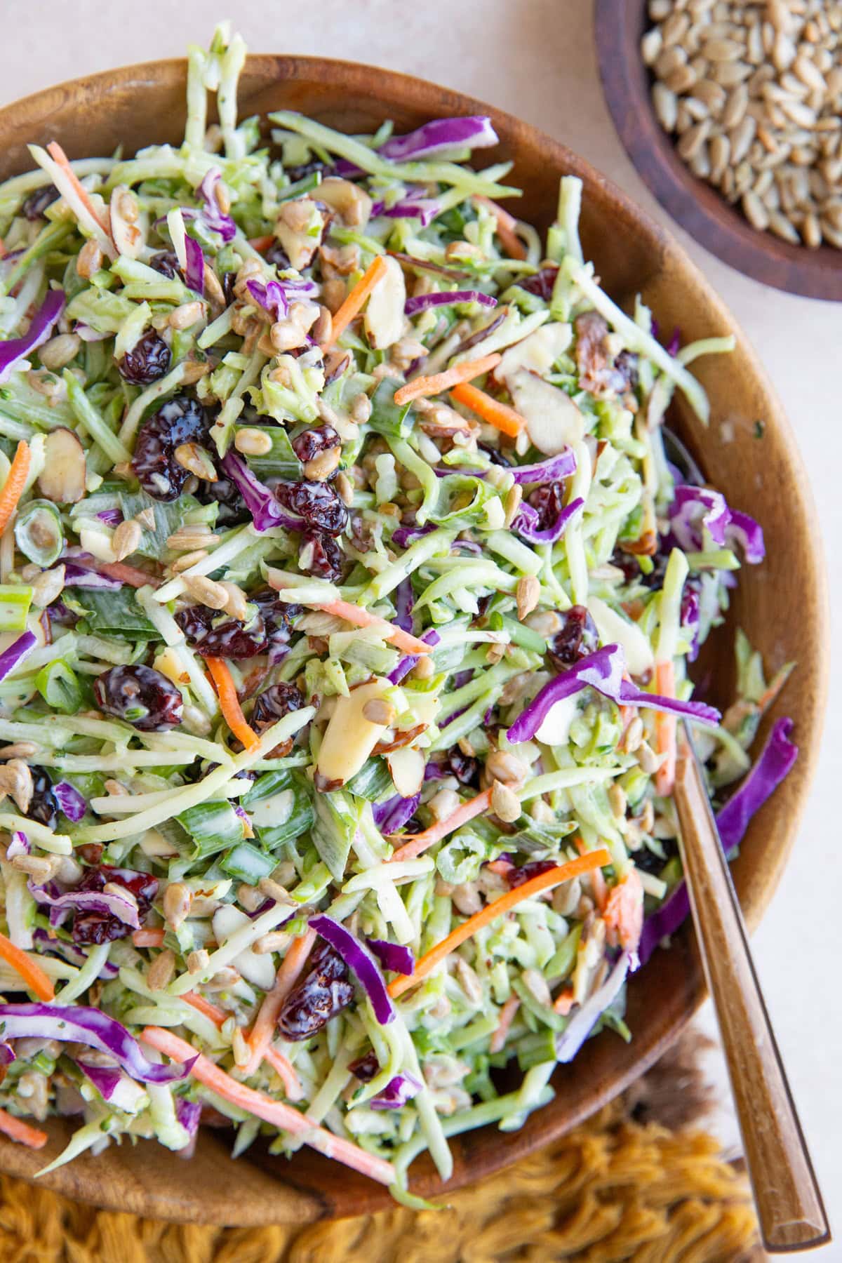 Wooden bowl full of broccoli slaw with a wood inlay spoon, ready to serve. A bowl of sunflower seeds to the side and a golden napkin.