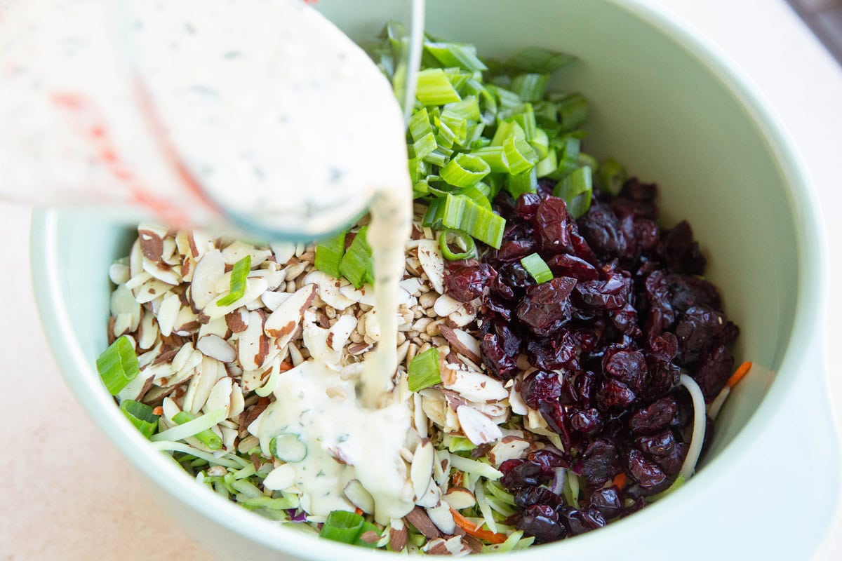 Pouring creamy coleslaw dressing into the bowl with the broccoli coleslaw ingredients.