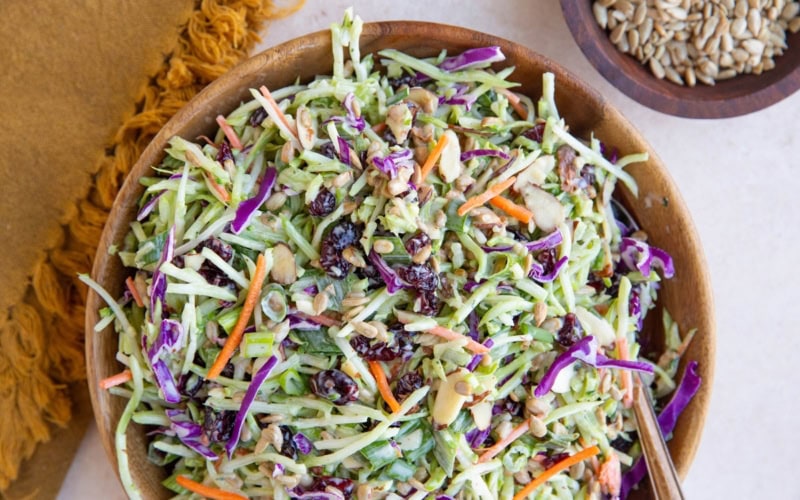 Broccoli coleslaw in a wooden bowl with a bowl of sunflower seeds and a golden napkin to the side. Ready to serve at any meal.