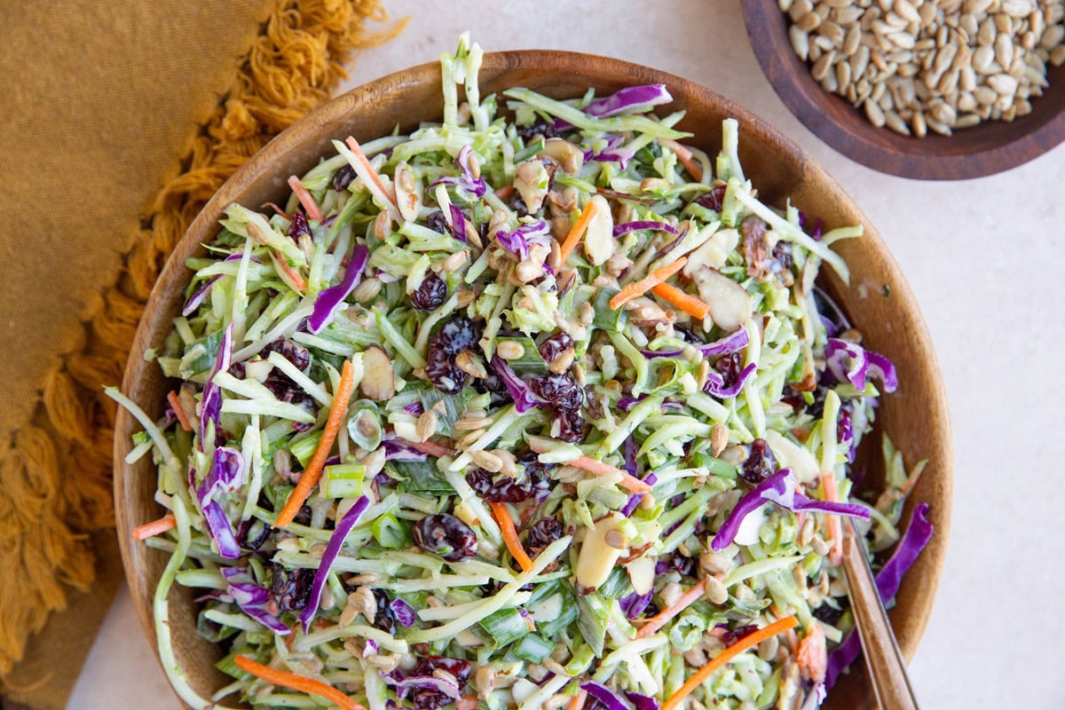 Broccoli coleslaw in a wooden bowl with a bowl of sunflower seeds and a golden napkin to the side. Ready to serve at any meal.