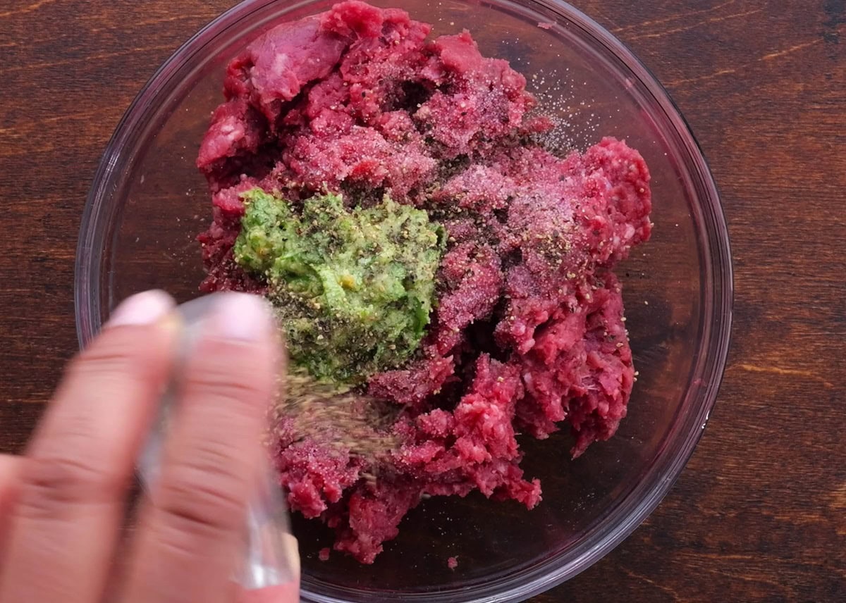 Ground beef and chimichurri sauce in a large bowl being sprinkled with salt and pepper.