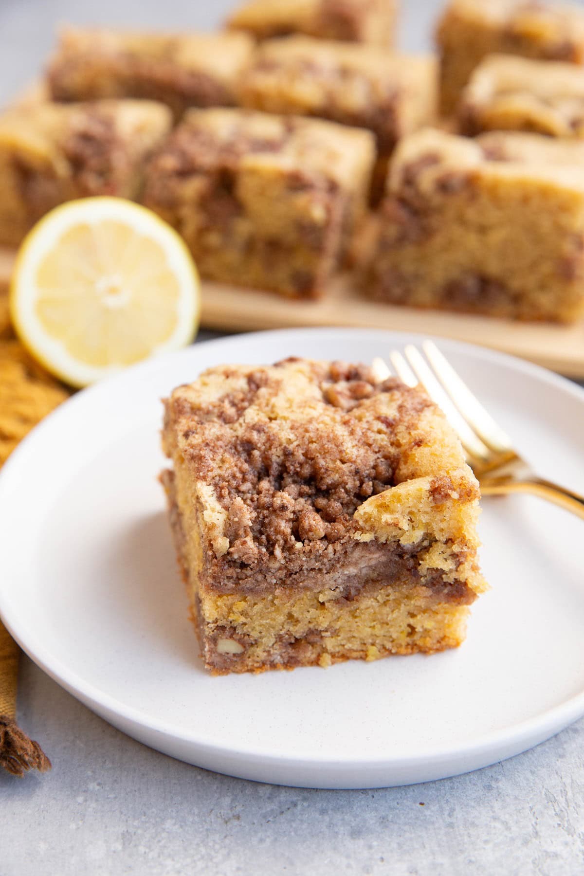 Big slice of lemon coffee cake on a white plate with a gold fork and the rest of the coffee cake in the background.