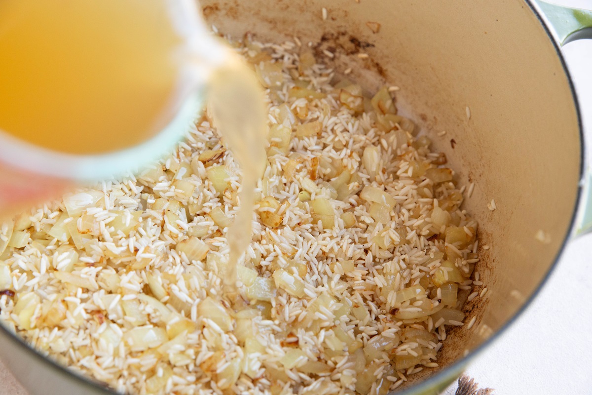 Pouring chicken broth into the pot with the rice and onions.
